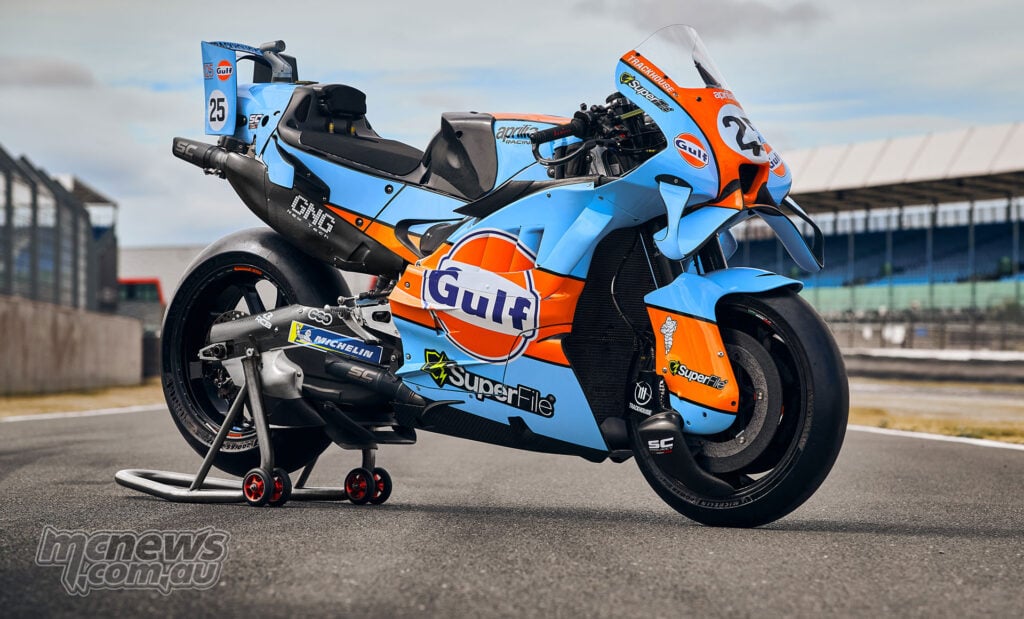 Three-quarter view of a Trackhouse Aprilia in light blue and orange Gulf livery, resting on a rear paddock stand.