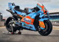 Three-quarter view of a Trackhouse Aprilia in light blue and orange Gulf livery, resting on a rear paddock stand.