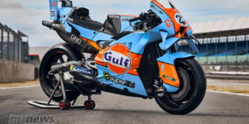 Three-quarter view of a Trackhouse Aprilia in light blue and orange Gulf livery, resting on a rear paddock stand.