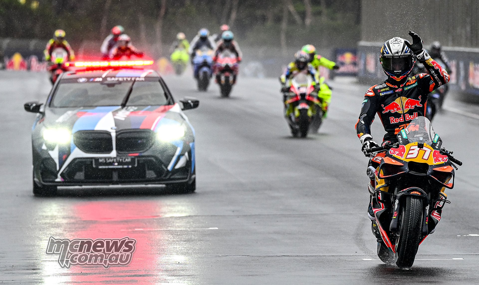 Pedro Acosta raises a hand from his number 37 KTM, with the BMW safety car and other riders on the wet Phillip Island straight.