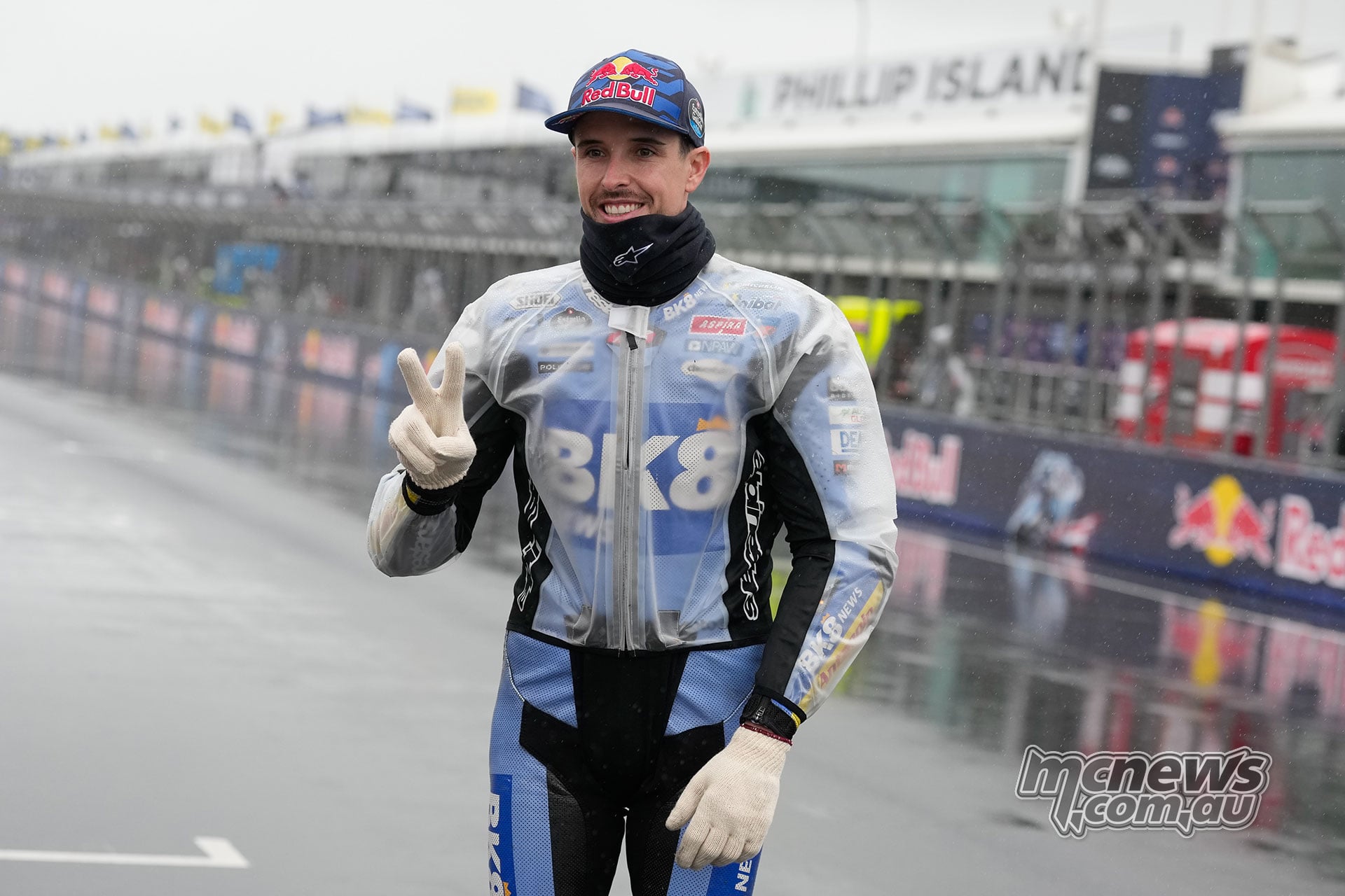 Alex Marquez holds up two gloved fingers while standing in Gresini leathers and a clear rain jacket on the wet grid.