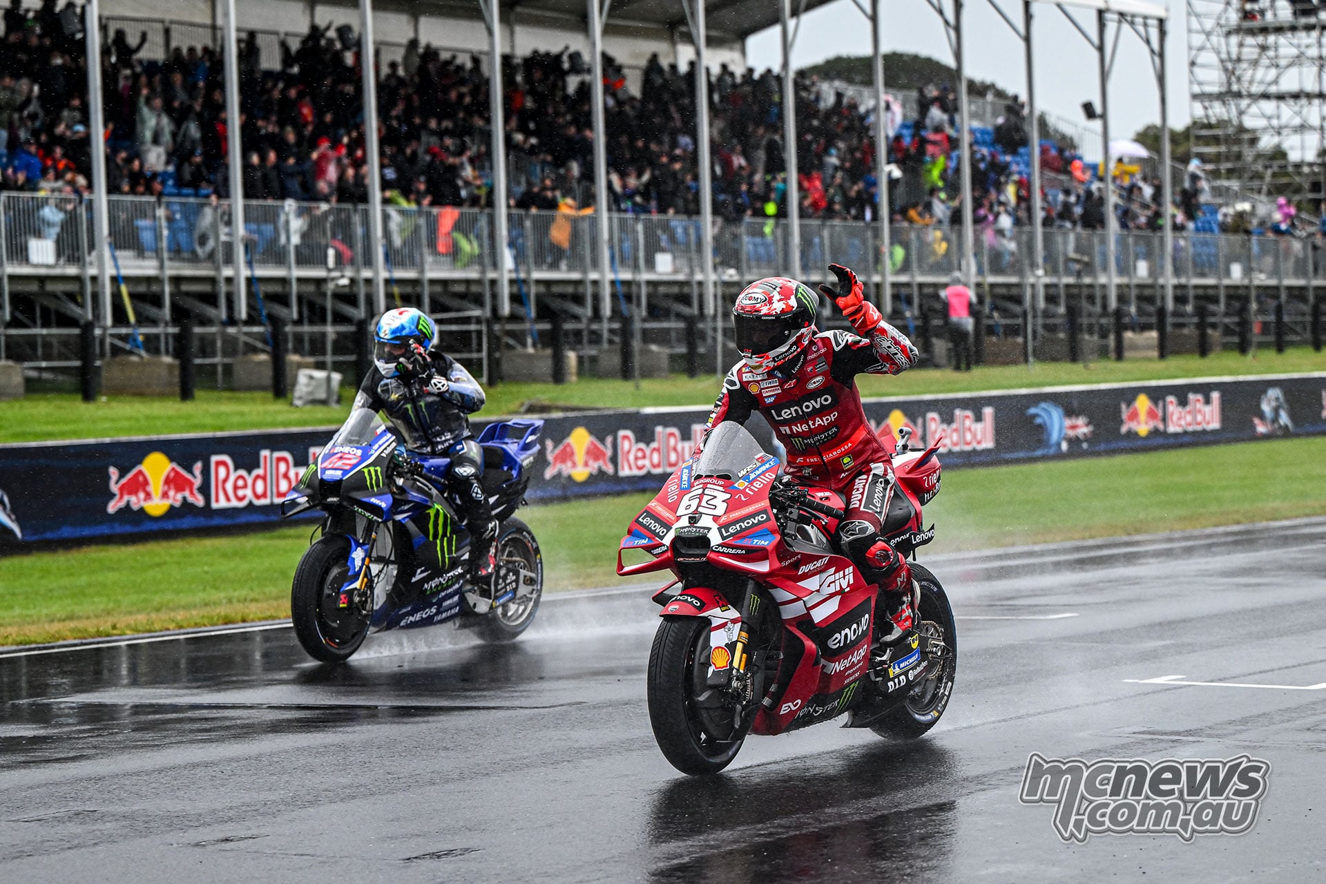 Alex Rins rides a blue Yamaha beside Francesco Bagnaia’s red number 63 Ducati as Bagnaia raises a hand towards the spectators.