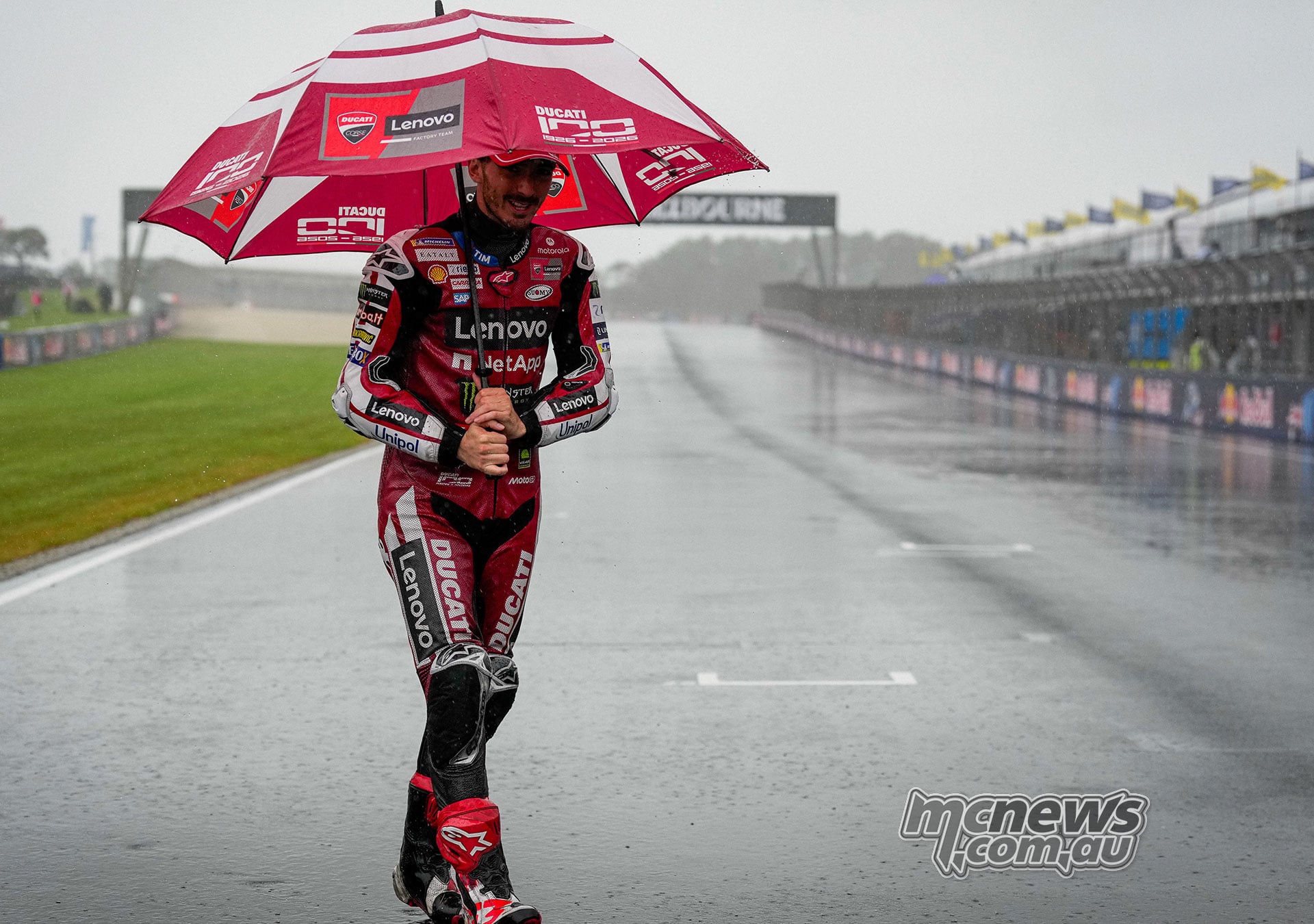 Francesco Bagnaia stands on the wet Phillip Island main straight holding a red Ducati Lenovo umbrella.