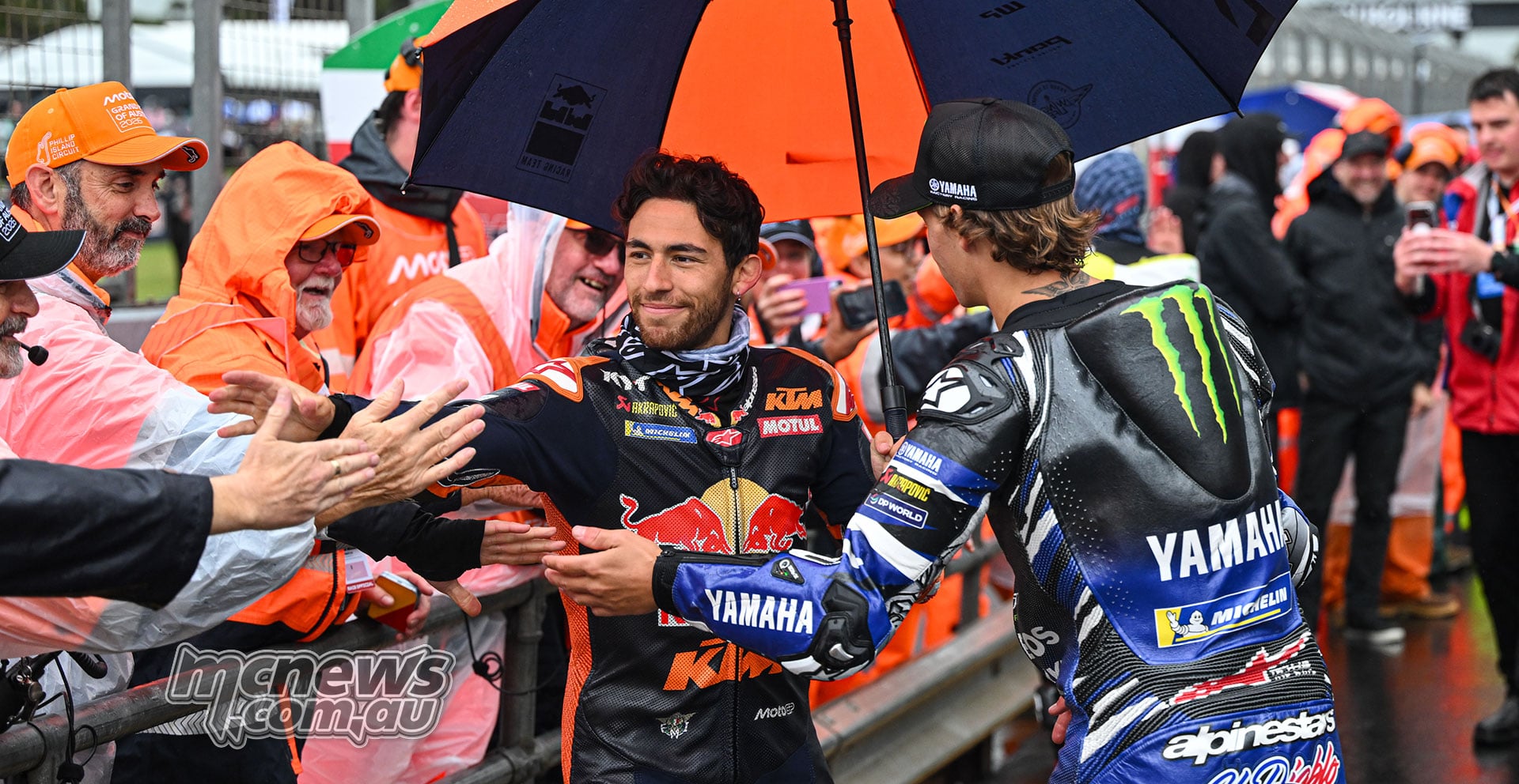 Enea Bastianini and Fabio Quartararo greet marshals in orange waterproof clothing beside the wet grid.
