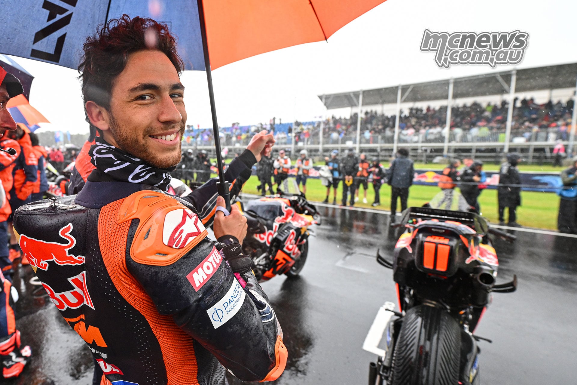 Enea Bastianini smiles beneath a KTM umbrella and gestures towards the grandstand, with KTM motorcycles behind him.