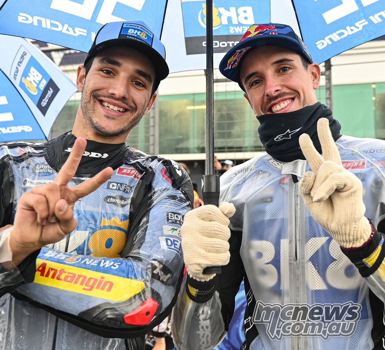 Iker Lecuona and Alex Marquez smile and make hand gestures beneath a blue-and-white Gresini Racing umbrella.