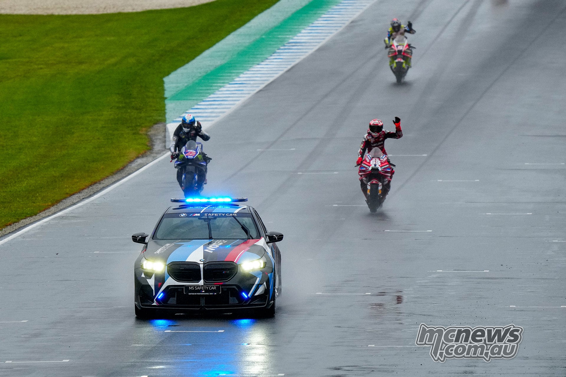 A BMW safety car with blue lights leads MotoGP riders along the wet main straight, with one rider waving.