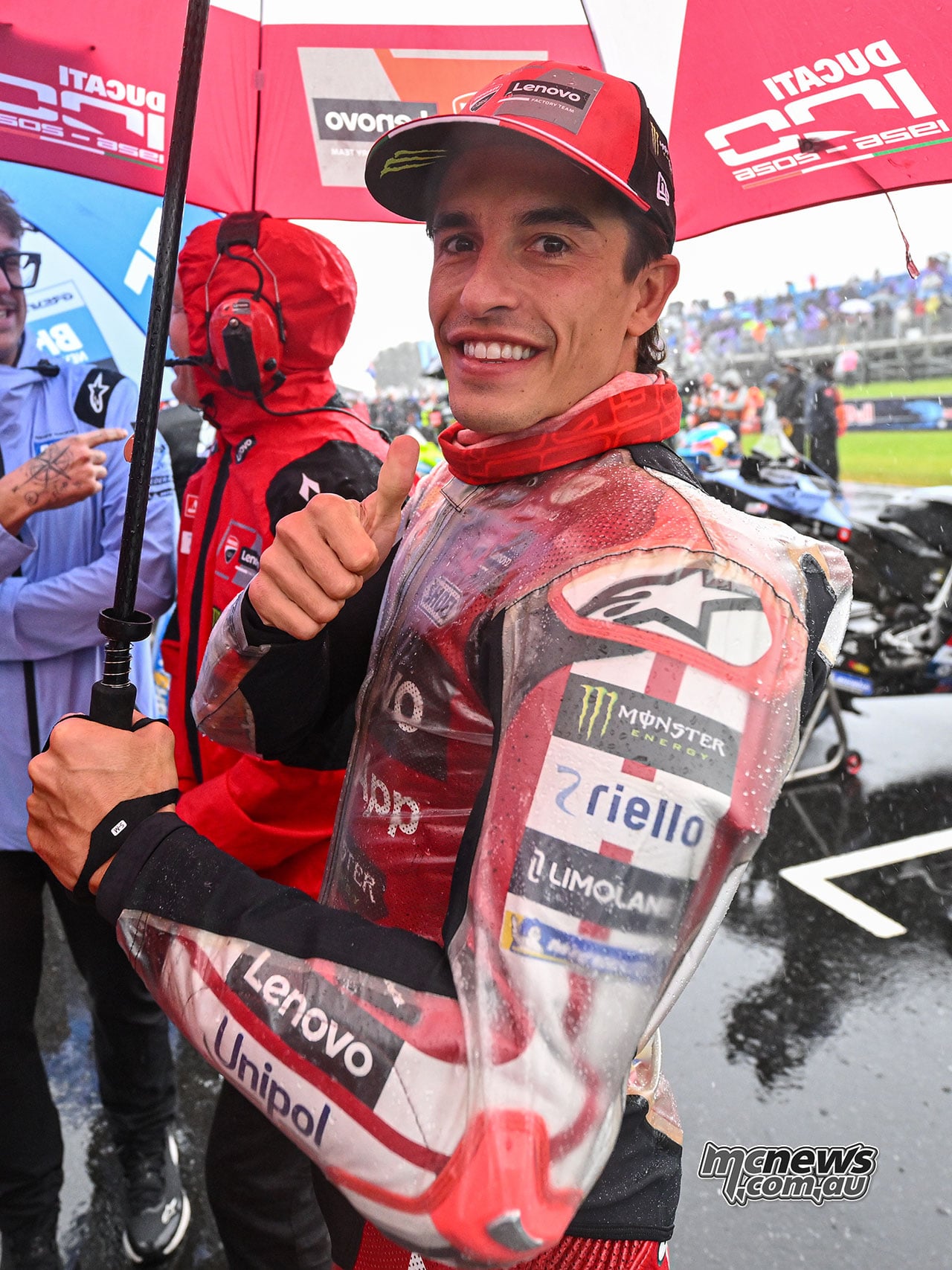 Marc Marquez holds a red Ducati umbrella and gives a thumbs-up while standing beside the motorcycles on the wet grid.