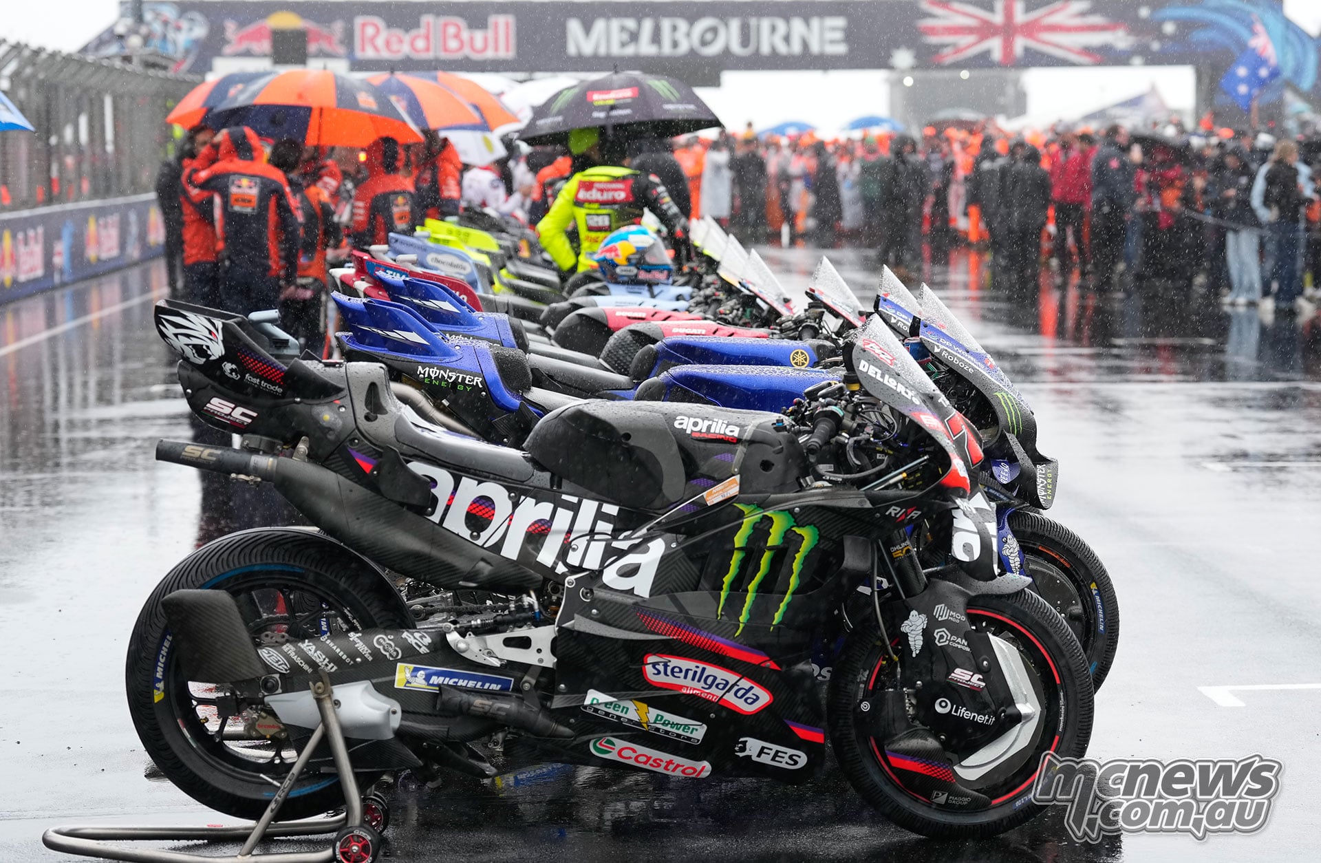 Rain-covered MotoGP motorcycles fitted with treaded tyres line the main straight, with an Aprilia in the foreground and people under umbrellas behind.