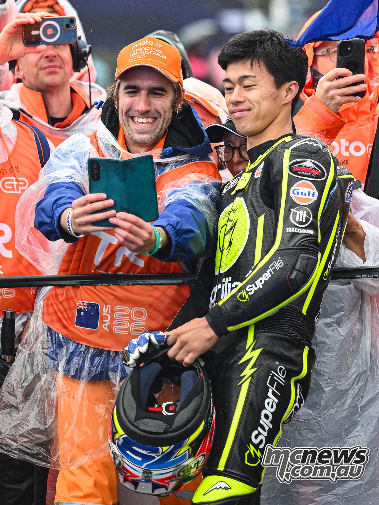 Ai Ogura poses beside a marshal holding a tablet, with other marshals photographing the moment behind them.