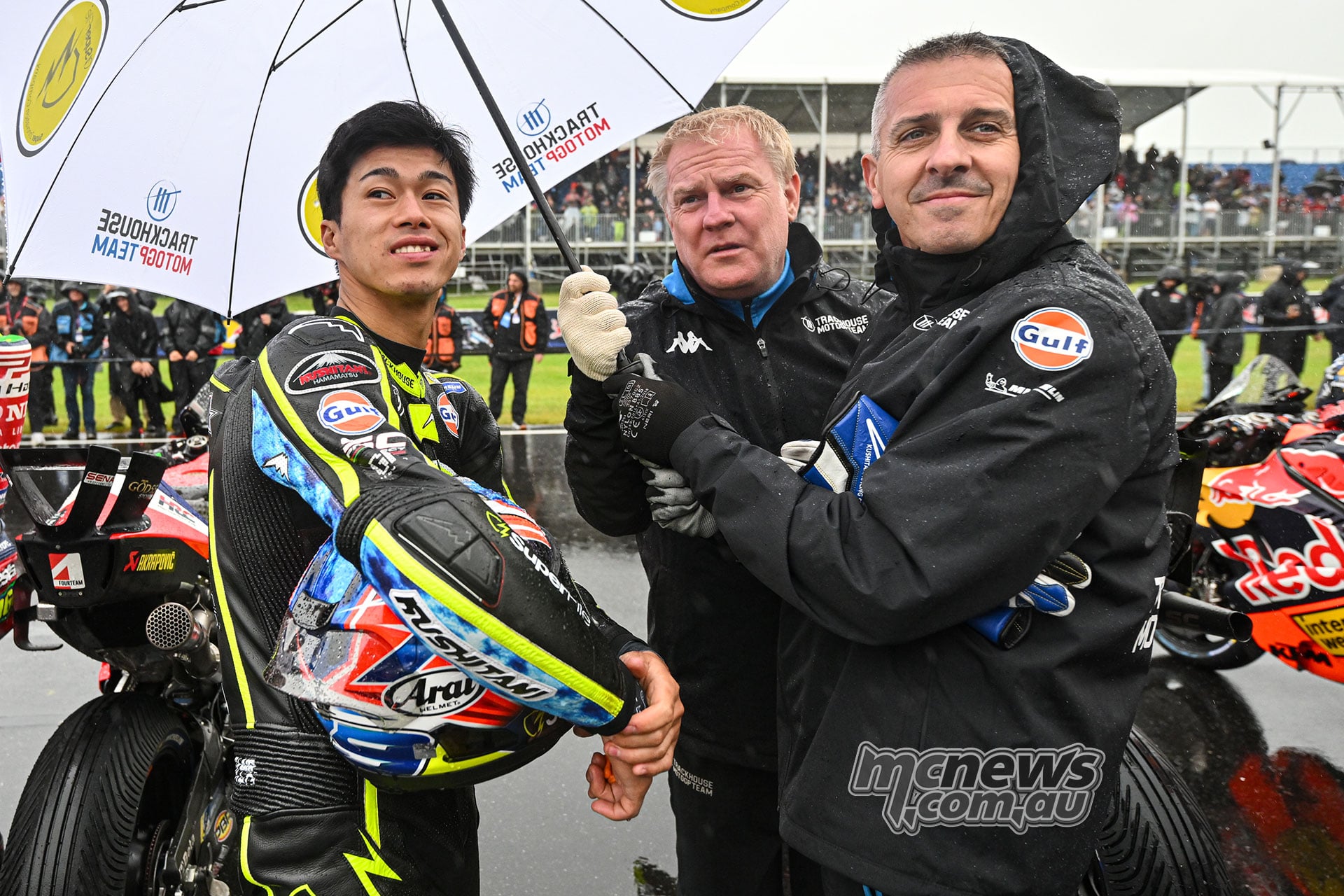 Ai Ogura holds his helmet beside two Trackhouse team members beneath a white umbrella on the wet grid.