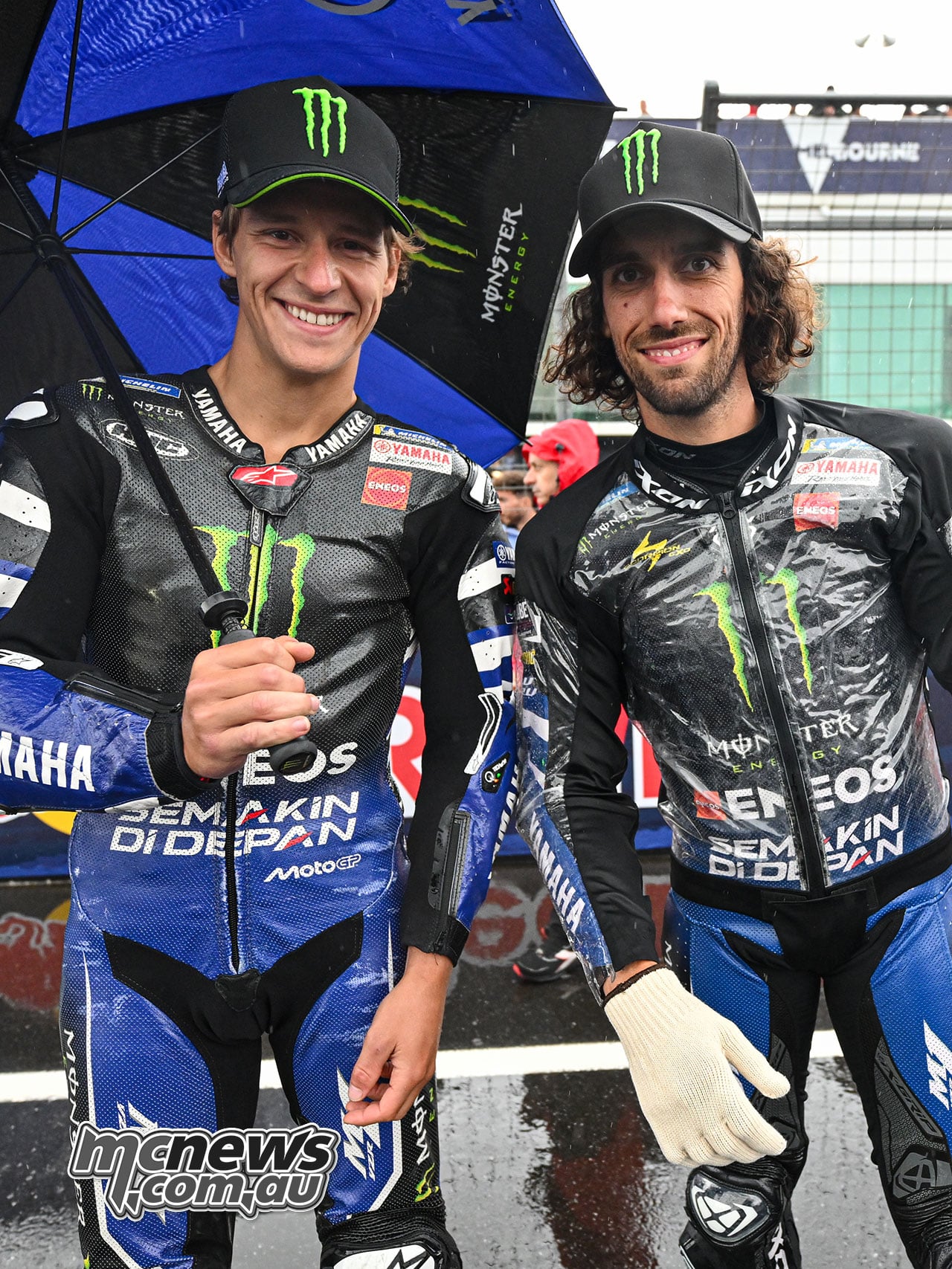 Fabio Quartararo and Alex Rins stand together beneath a blue-and-black Monster Energy Yamaha umbrella on the wet grid.