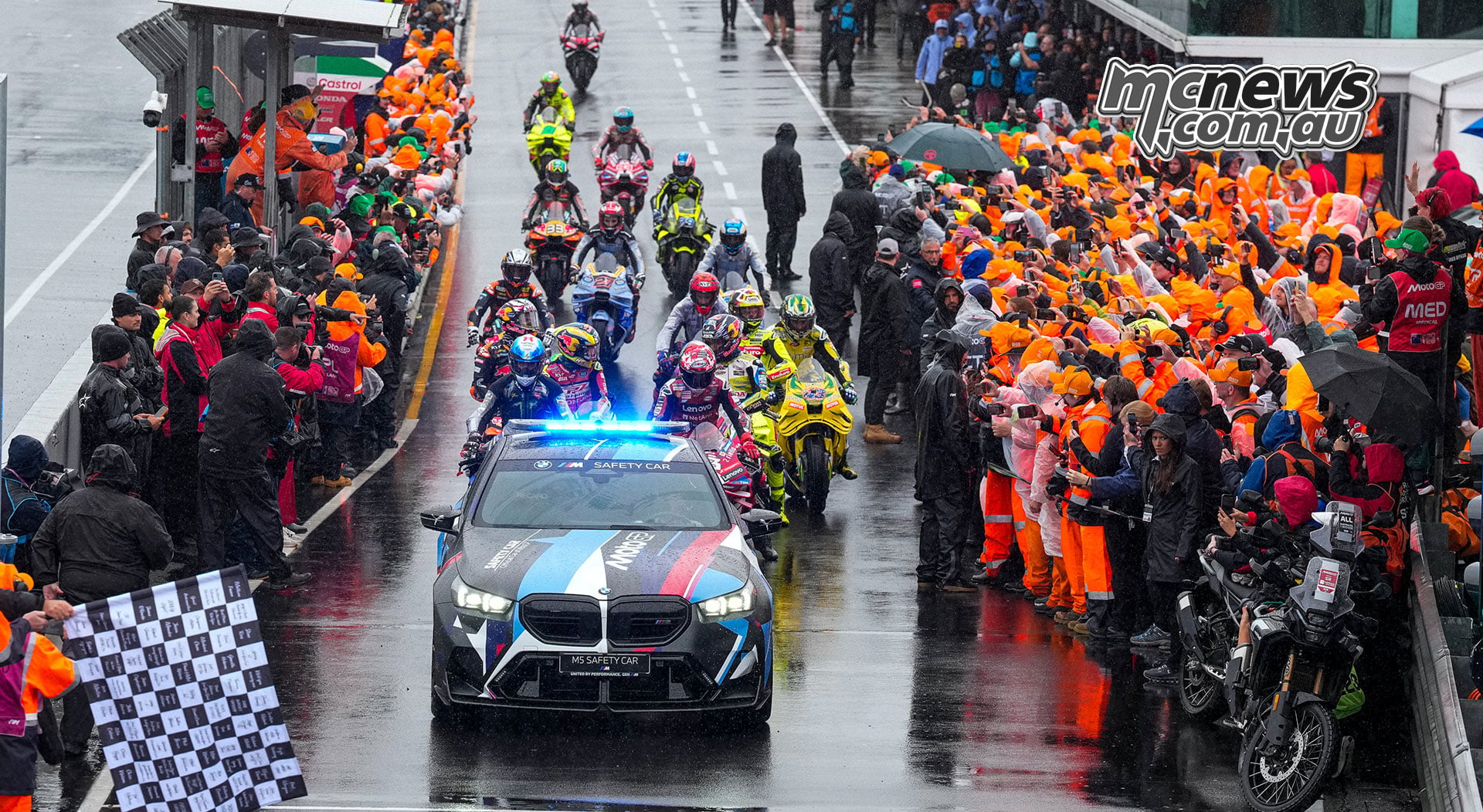 The BMW safety car and MotoGP riders pass between marshals in orange and team personnel lining the wet pit lane.
