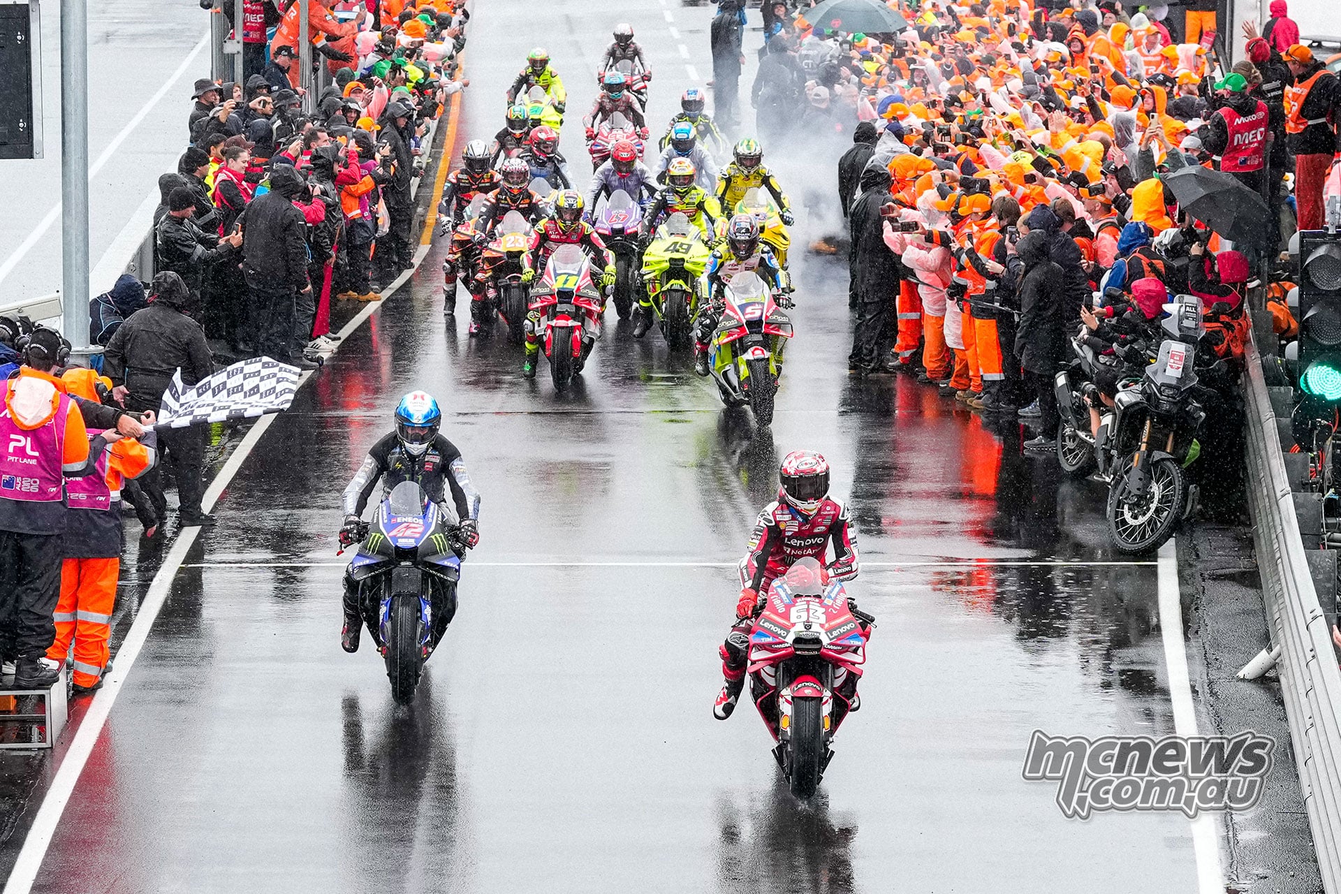 MotoGP riders leave the wet pit lane between lines of marshals and team personnel, with a chequered flag at the side.