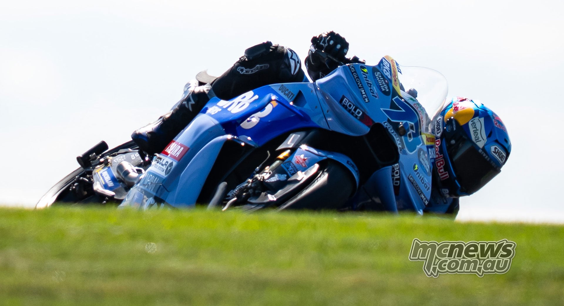 Alex Marquez leans his blue number 73 Gresini Ducati deeply into a corner, photographed from behind a grassy rise.