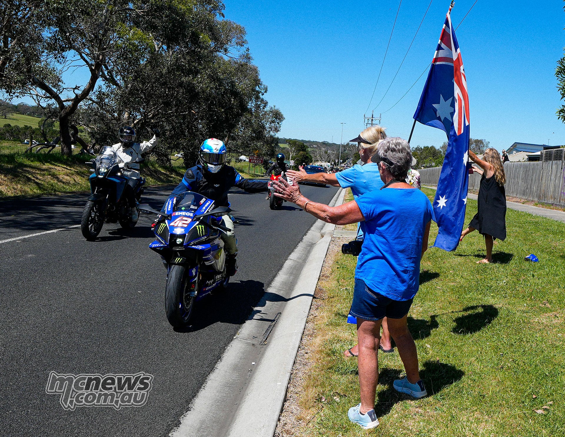 Alex Rins reaches towards spectators for a high-five while riding a blue number 42 Yamaha on a public road beside an Australian flag.