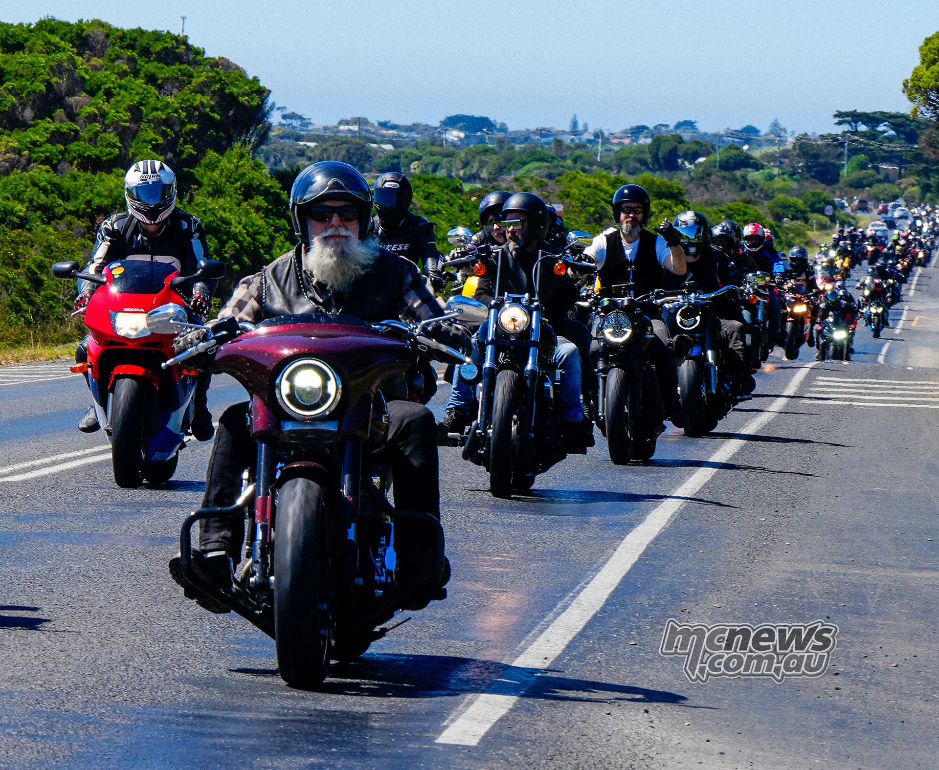 A long convoy of motorcyclists rides along a sunny road, led in the foreground by a rider on a maroon touring motorcycle.