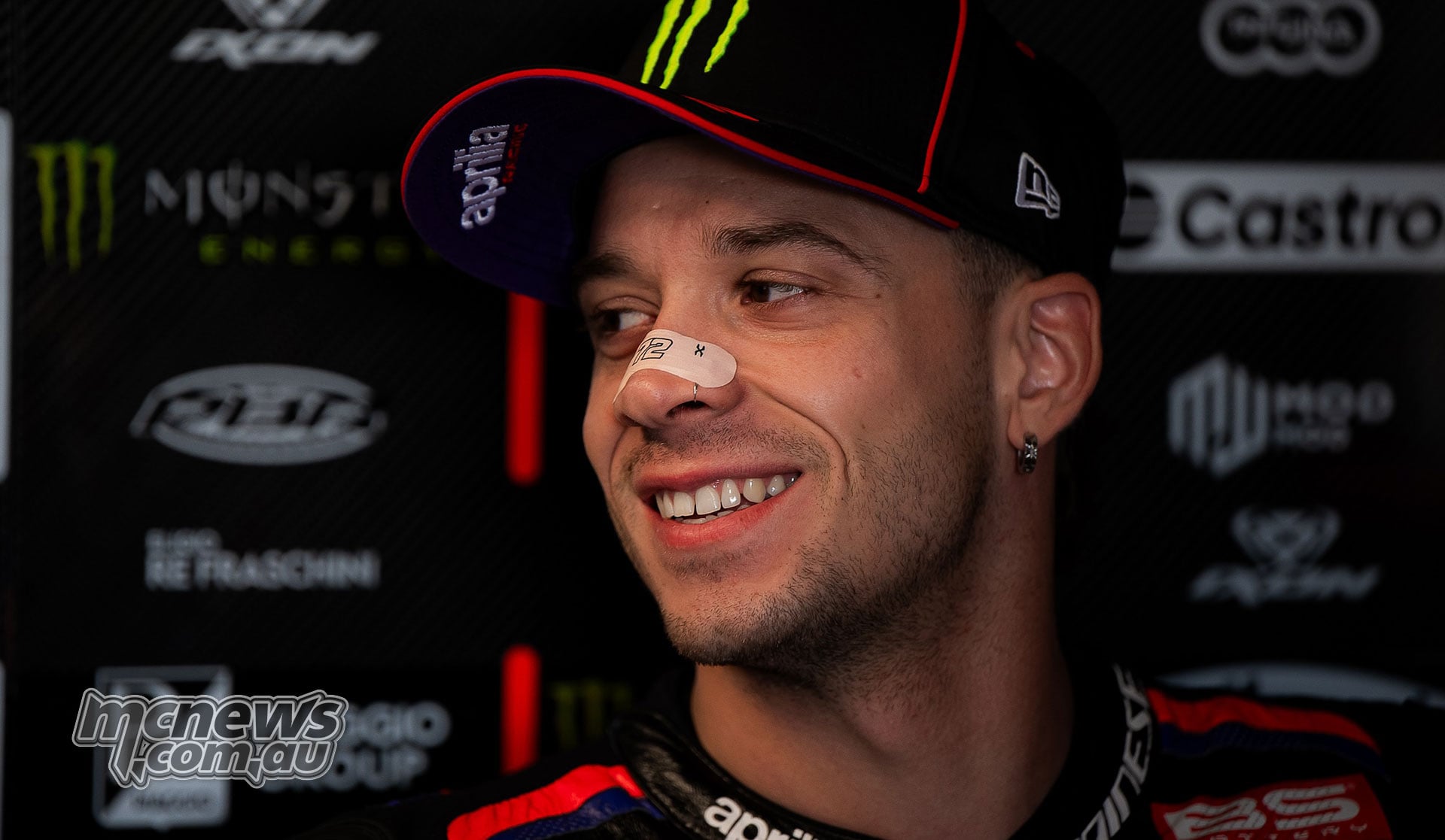 Marco Bezzecchi smiles in the Aprilia garage wearing a team cap and a nasal strip.