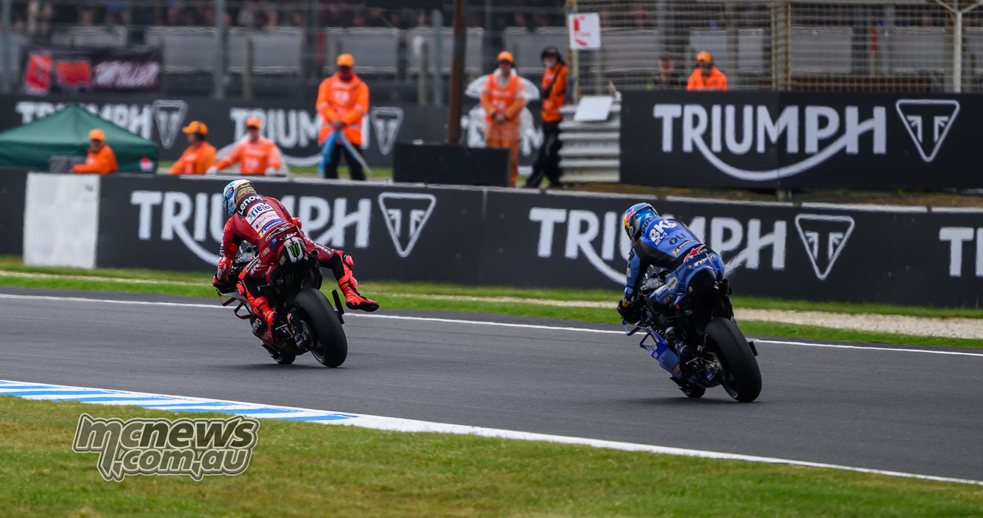 Marc Marquez leads Alex Marquez towards a corner at Phillip Island, both viewed from behind with marshals beside the track.