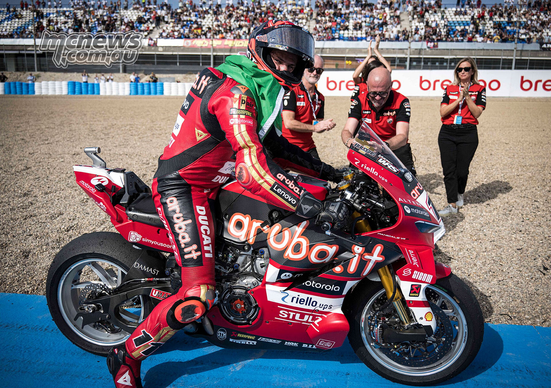 Nicolò Bulega celebrates his fiftieth WorldSBK win with the Italian flag draped over his shoulders aboard his Ducati as team members applaud at Jerez