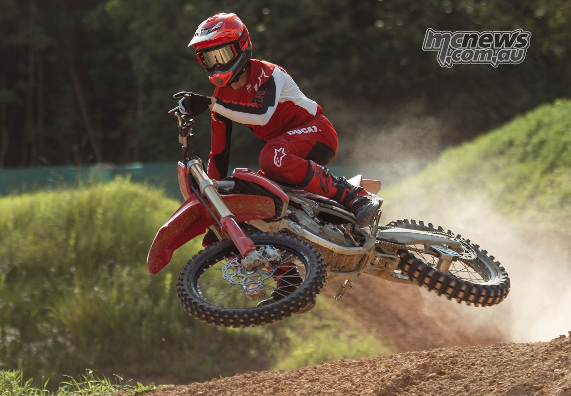 Motocross rider in red and white Ducati racing gear whipping a red Desmo450 MX sideways through the air above a dirt jump.