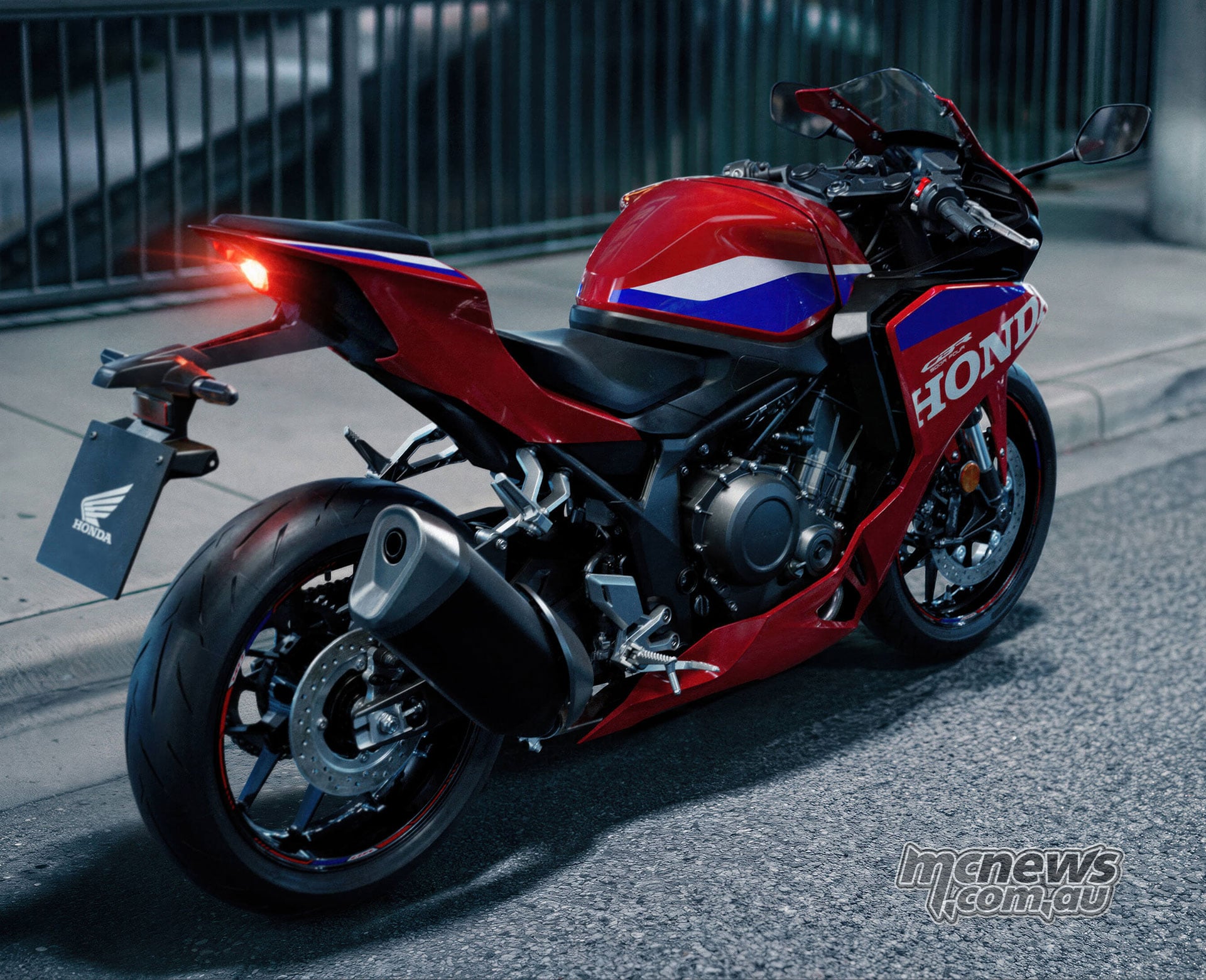 Rear right three-quarter view of a red 2027 Honda CBR500R FOUR parked on a city street with its tail light illuminated