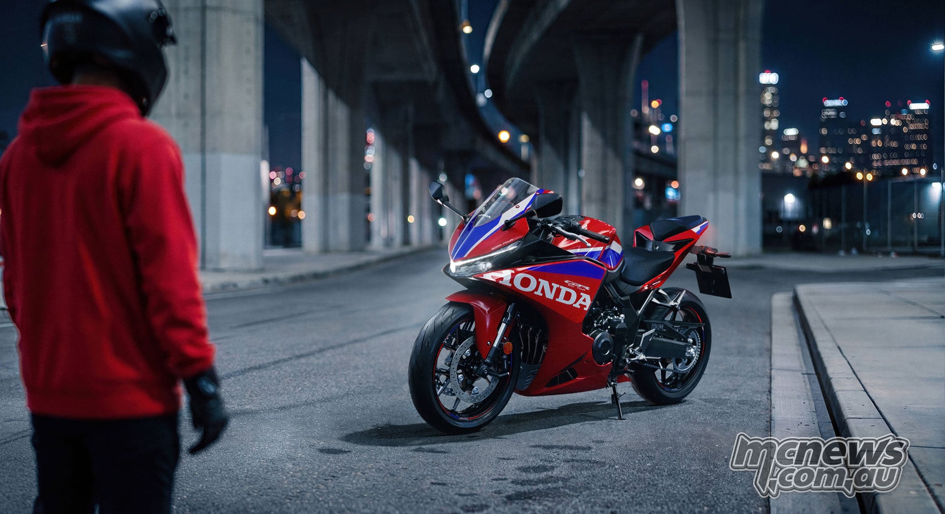 Helmeted rider in a red hoodie facing a parked 2027 Honda CBR500R FOUR beneath an elevated roadway at night