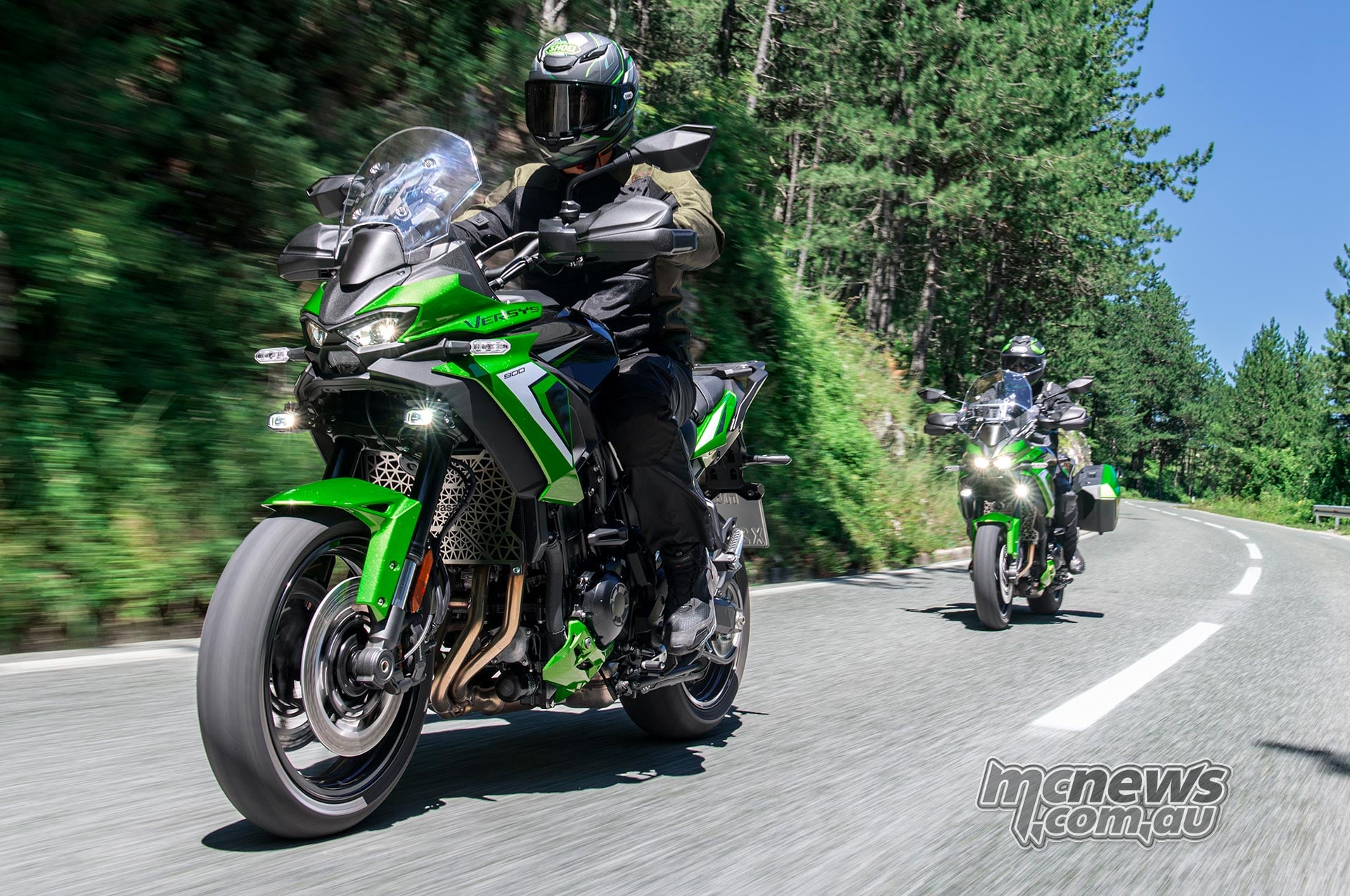 Two riders on green Kawasaki Versys 900 SE motorcycles travelling along a winding tree-lined road