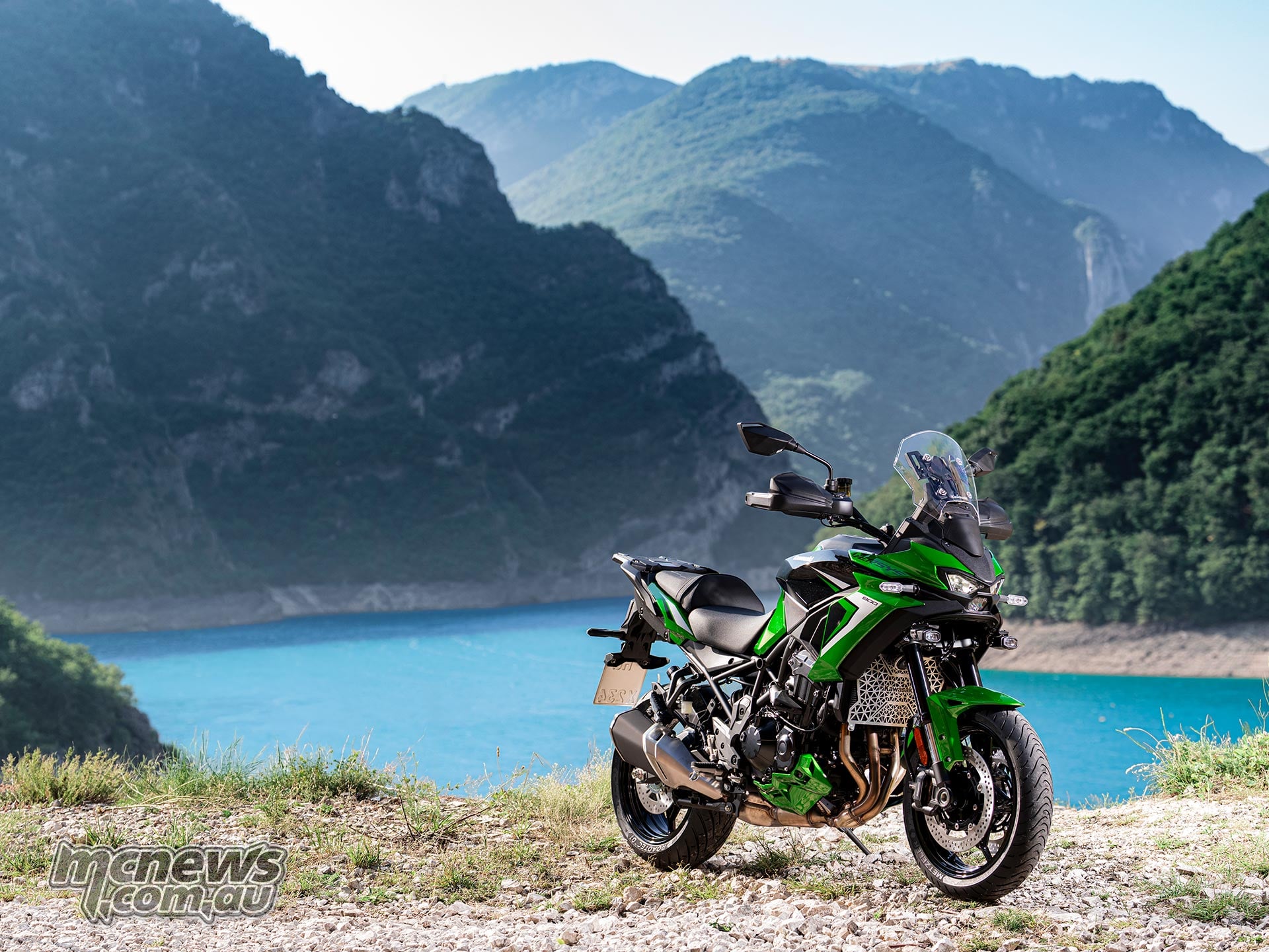 Green Kawasaki Versys 900 SE parked beside a turquoise lake with mountains in the background