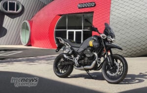 The V85 Strada wears the pearlescent grey and yellow anniversary livery inspired by the redeveloped Mandello complex. Grey and yellow Moto Guzzi V85 Strada anniversary edition parked in front of the red shop entrance at Mandello.