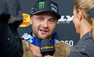 Cooper Webb speaks to a television interviewer with mud on his jersey and goggles around his neck.