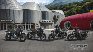 From left: Stelvio, V100 Mandello, V85 Strada and V7 Stone in 105th-anniversary colours outside Moto Guzzi’s new museum in Mandello del Lario. Four grey Moto Guzzi motorcycles with yellow anniversary graphics outside the new museum’s sculpted metal buildings, with mountains behind.