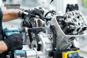 Modern production still has a human touch, with a worker securing an alternator during V-twin engine assembly. Gloved hands secure an alternator on a Moto Guzzi V-twin engine with its rocker cover removed.