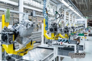 Engines line up at Mandello, where Piaggio quotes annual production capacity of more than 30,000 motorcycles following the redevelopment. Rows of Moto Guzzi V-twin engines mounted on yellow carriers along the bright factory assembly line.