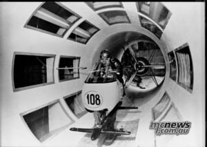 Rider and motorcycle under examination in Moto Guzzi’s wind tunnel in the 1950s, when streamlining became a crucial part of Grand Prix development. Black-and-white photograph of a rider crouched on a number 108 motorcycle inside Moto Guzzi’s wind tunnel.
