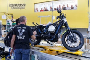 A window onto the present: museum visitors can look straight into the factory where new Moto Guzzis take shape. Partly assembled Moto Guzzi on a yellow production carrier, with visitors watching through the window behind.