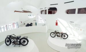 Early machines occupy a dramatically different home inside the new 3000-square-metre Moto Guzzi museum. Visitors lean in and take photographs of an early olive-green Moto Guzzi displayed on a white museum plinth.